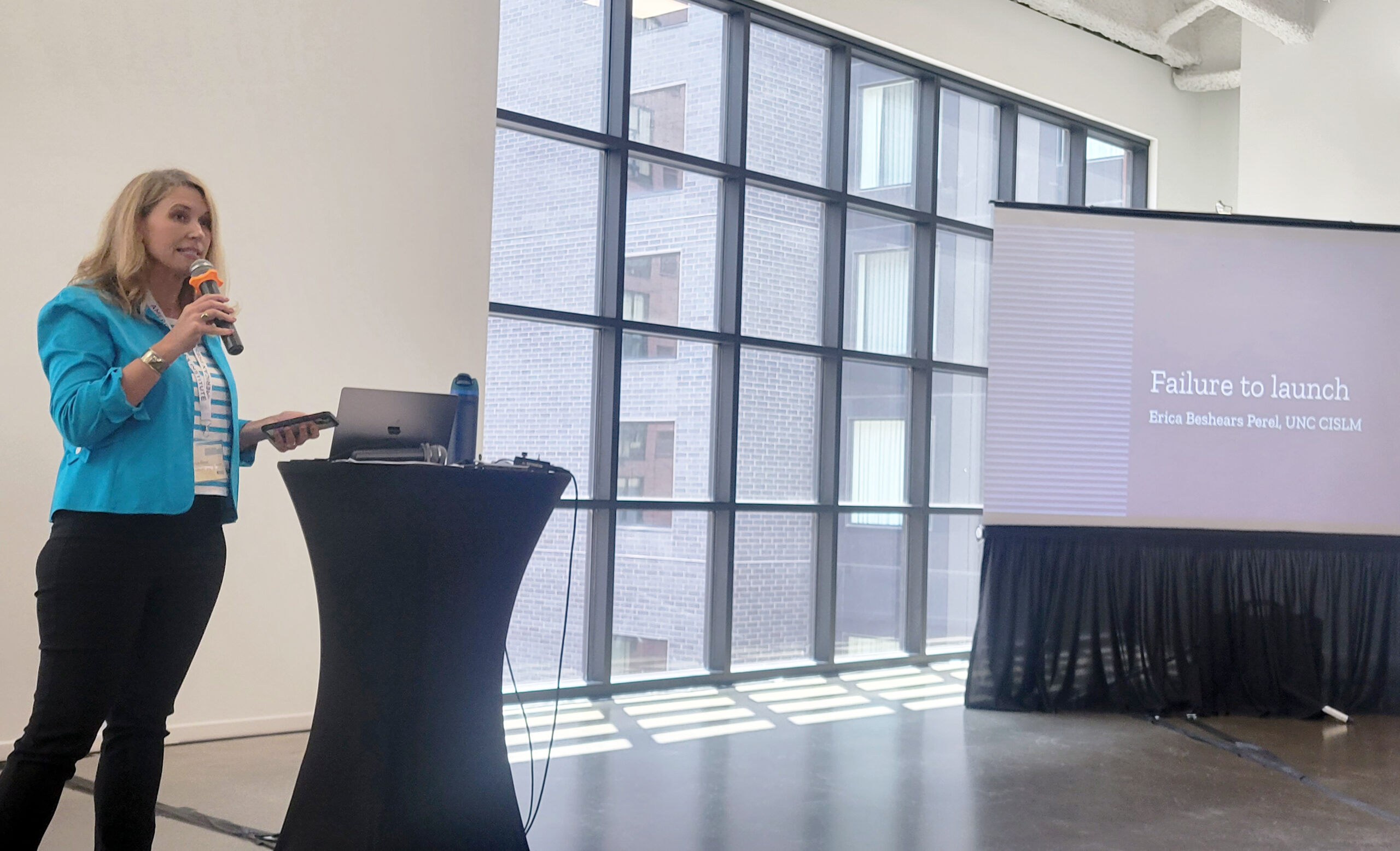 A woman stands beside a table and speaks into a microphone. A screen to her left shows the title of her talk, "Failure to launch"