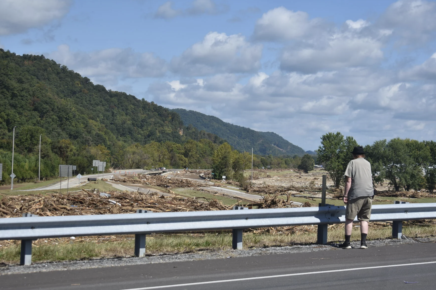 A man stands on the side of the road looking at flood debris that stretches from the side of the road onward