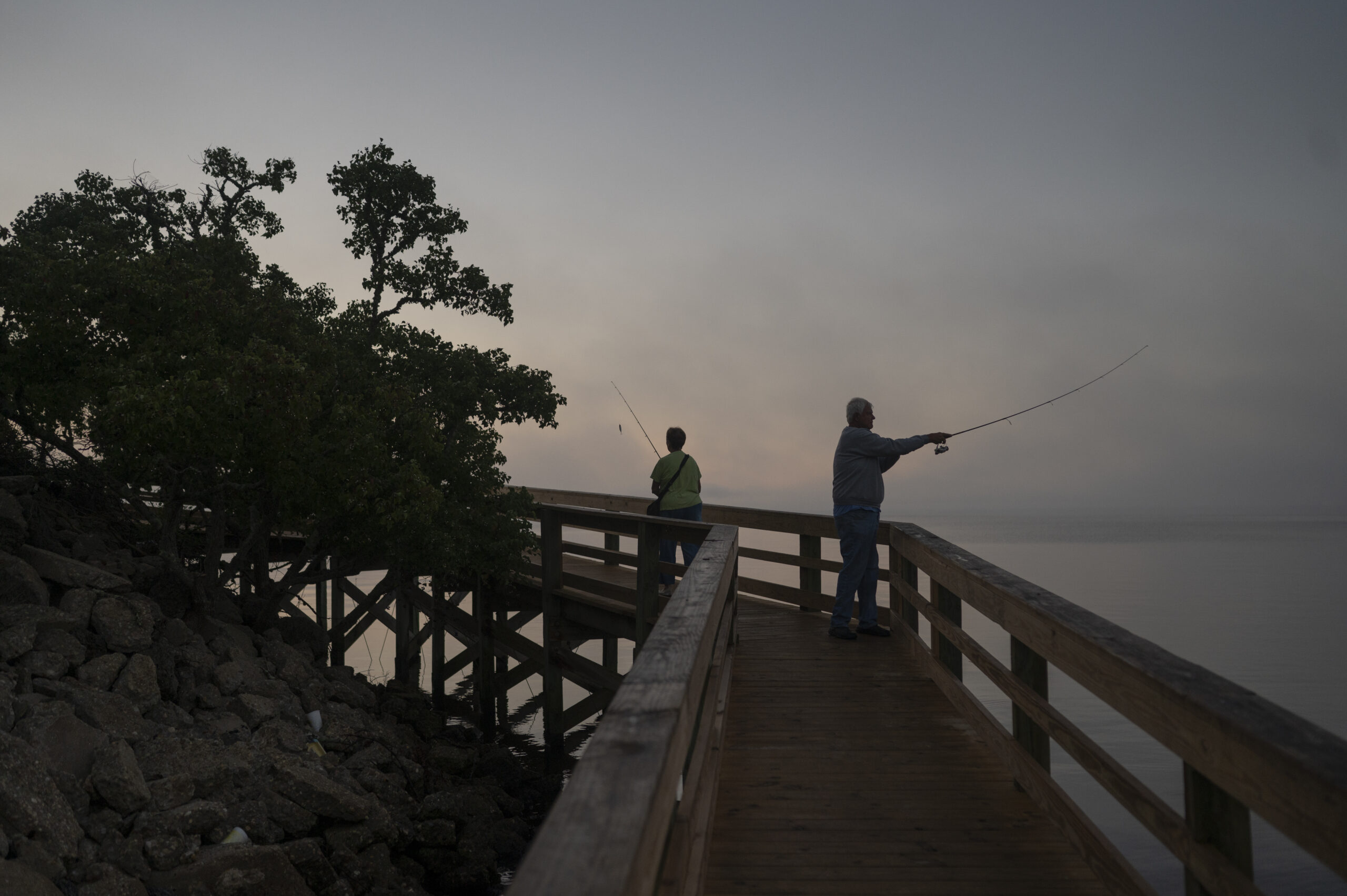 A woman and man cast lines into a river from a pier in the early dawn light