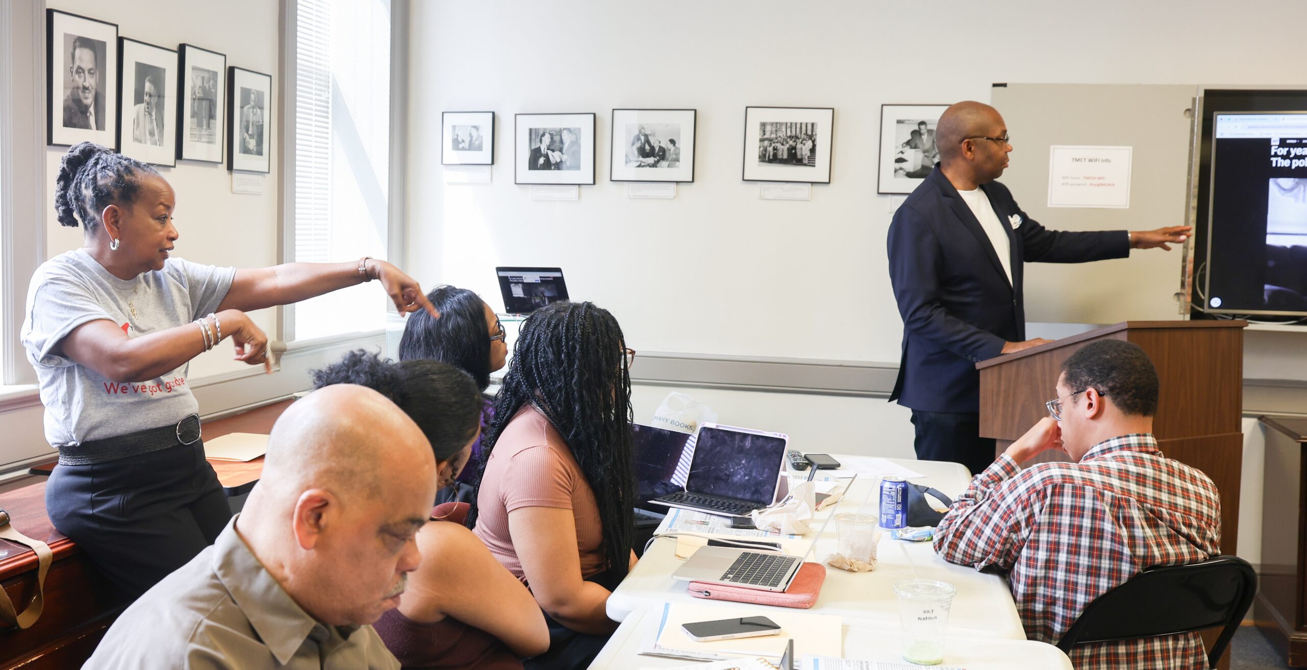 A lively journalism workshop in session: a woman gestures while engaging attendees, who are seated with laptops and notes. A presenter points to a screen at the front of the room. Framed photos and large windows line the walls of the learning space.
